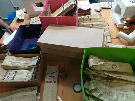 Was auf den ersten Blick etwas chaotisch aussieht, ist ein gut sortierter Arbeitsplatz. Auf der linken Platzseite werden die Kaffeepackungen schon mal fertig zum Vernähen gemacht. Und auf der rechten Seite werden die bereits vorbereiteten Packungen mit der Nähmaschine vernäht, so entsteht Schritt für Schritt eine Tasche aus Kaffeepackungen.